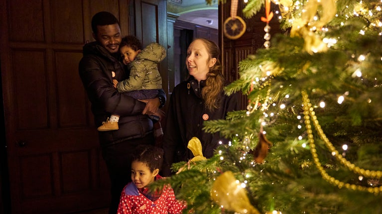 A family with two adults and two children stands in the doorway of a room with dark wooden panelling, admiring the large Christmas tree lit up in the foreground of the image.